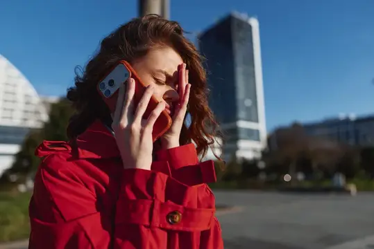 Молодая женщина в ярко-красном пальто прижимает смартфон к уху и частично закрывает лицо рукой, проявляя признаки стресса во время телефонного разговора в городской обстановке с высокими зданиями и