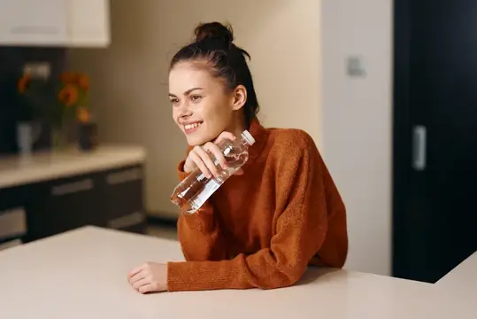 Улыбающаяся девушка с бутылкой воды за столом на кухне