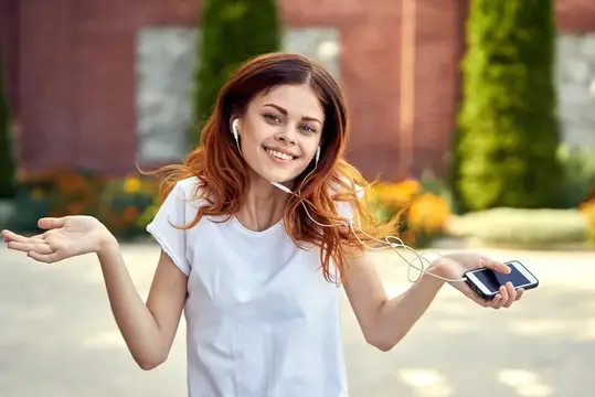 Рыжеволосая девушка в наушниках слушает музыку с телефона на улице