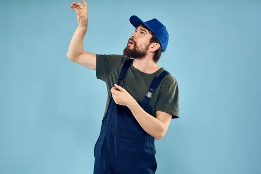 Бородатый мужчина в синем комбинезоне и кепке смотрит вверх на голубом фоне