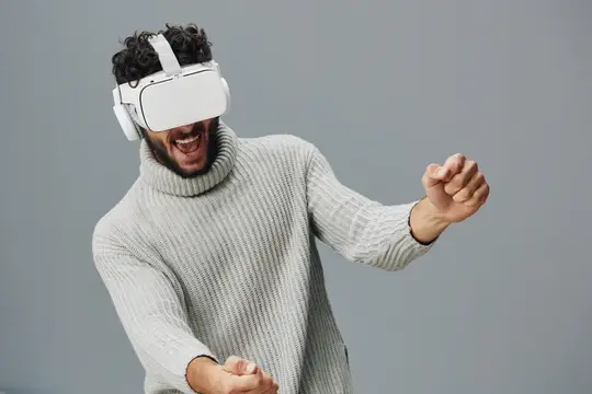 Кудрявый мужчина в VR-очках радостно играет в светлой вязаной кофте