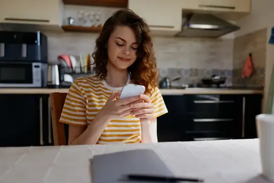 Кудрявая девушка в полосатой футболке листает телефон на кухне