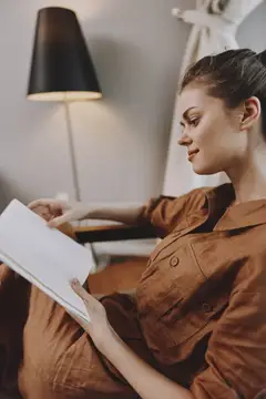 Молодая женщина в коричневом платье читает книгу в кресле