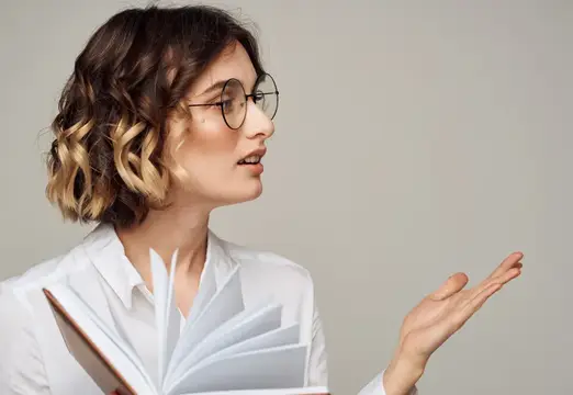 Эмоциональная женщина жестикулирует руками на сером фоне, на лице очки, прическа для образования