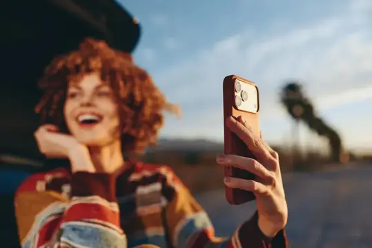 Рыжеволосая девушка со смехом фотографирует на смартфон на закате