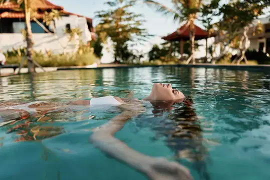 Лайфстайл-сцена спокойного отпуска у чистого бассейна: фигура, плавающая на поверхности, в залитом солнцем тропическом курорте с пальмами, водой и расслабленным настроением