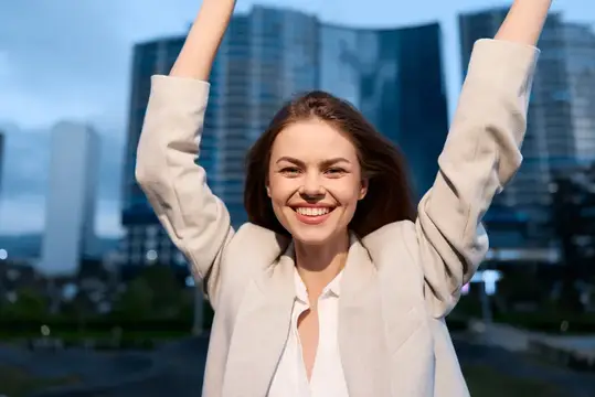 Радостная деловая женщина с яркой улыбкой поднимает руки в жесте победы на фоне современного горизонта, символизируя достижение и амбиции в своей карьере