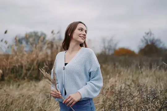 Улыбающаяся девушка в голубом кардигане стоит в осеннем поле