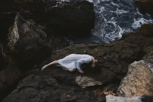 Женщина в белом платье лежит на тёмных прибрежных камнях у бурного моря