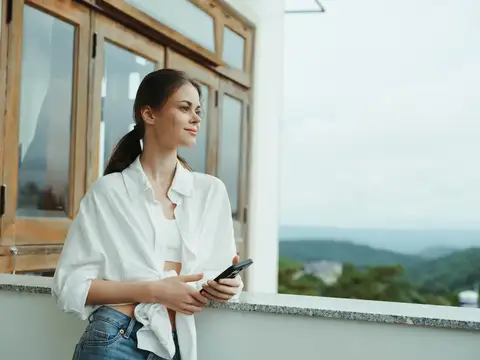 Женщина на балконе дома с телефоном, красивая улыбка в выходной день, прекрасный вид на природу в горах, образ жизни и отдых в отеле