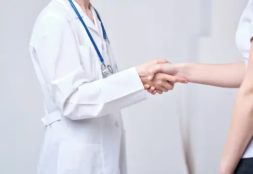 Врач и пациентка пожимают друг другу руки на светлом фоне, кадр обрезан