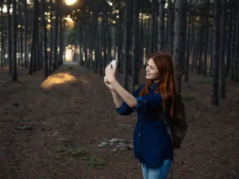 Рыжеволосая девушка фотографирует на телефон в сосновом лесу