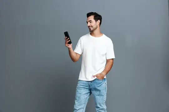 Улыбающийся бородатый мужчина в белой футболке смотрит на телефон на сером фоне