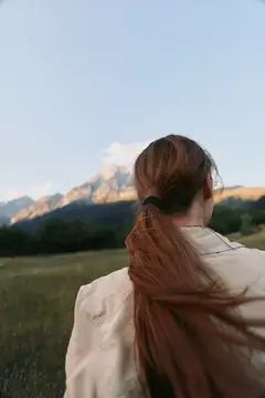 Вид сзади на молодую женщину с хвостиком, одетую в легкую куртку, стоящую на травянистом поле и смотрящую на далекие горы под голубым небом, передающий образ жизни и природу