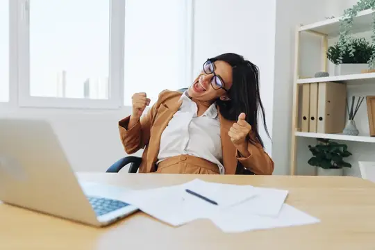 Деловая женщина работает в офисе за столом с ноутбуком, показывает жест победы и радуется успеху