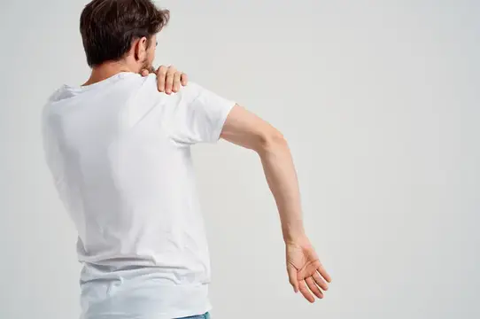Мужчина в белой футболке массирует ноющее плечо на светлом фоне