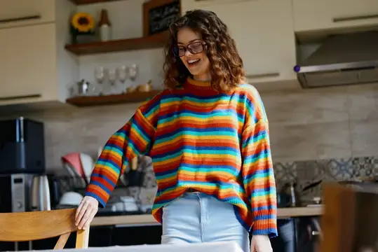 Улыбающаяся кудрявая девушка в очках и радужном свитере на кухне