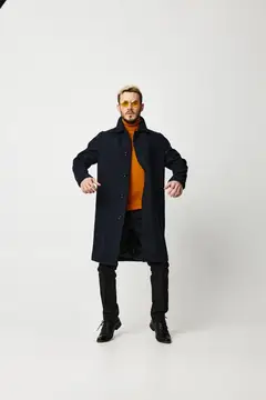 Модный мужчина в пальто держит руки на поясе, на нем черные брюки, модель на светлом фоне