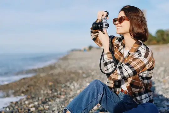 Улыбающаяся девушка в клетчатой рубашке фотографирует на камеру у моря