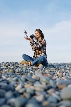 Девушка с фотокамерой сидит на каменистом пляже и подбрасывает камешки