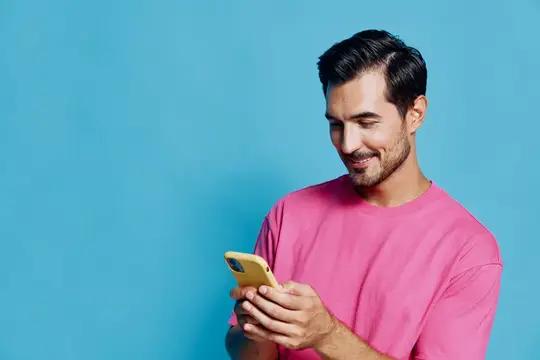 Улыбающийся брюнет в розовой футболке смотрит в смартфон на синем фоне