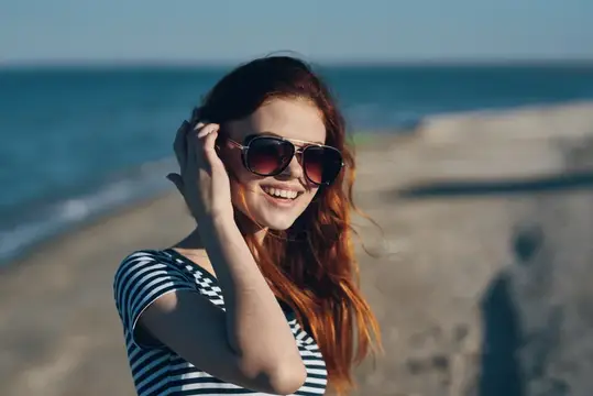 Рыжеволосая девушка в солнечных очках улыбается на пляже у моря