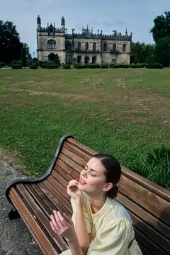 Молодая женщина с закрытыми глазами сидит на скамейке в парке у старинного замка