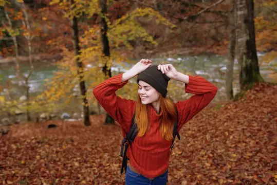 Женщина-туристка в свитере и шапке с рюкзаком стоит среди высоких деревьев в осеннем лесу