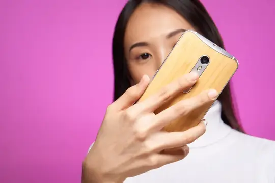 Азиатская девушка закрывает лицо смартфоном на розовом фоне