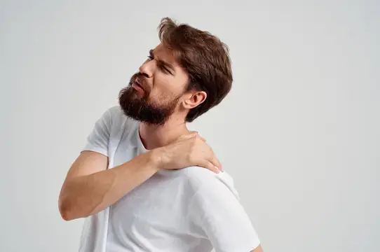 Бородатый мужчина в белой футболке массирует ноющее плечо