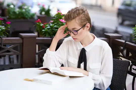 Молодая женщина в очках читает книгу за столиком летнего кафе