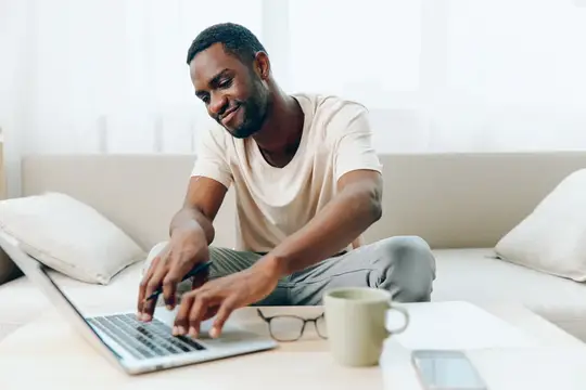 Улыбающийся темнокожий мужчина работает за ноутбуком на диване