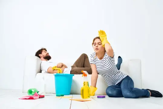 Уставшая женщина в перчатках убирается, пока мужчина отдыхает на диване