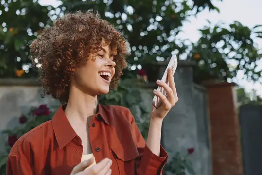 Кудрявая девушка в рубашке смеётся, глядя на смартфон в саду