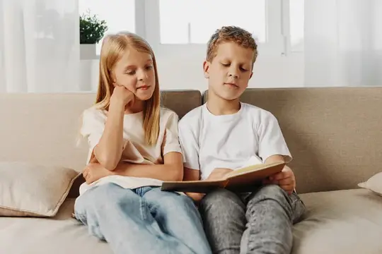 Белокурая девочка и мальчик читают книгу на диване