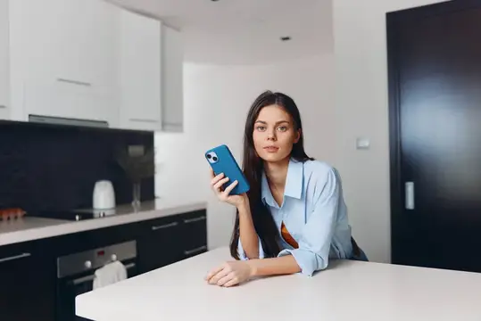 Молодая женщина сидит на кухонной стойке, держит в руках мобильный телефон и смотрит прямо в камеру