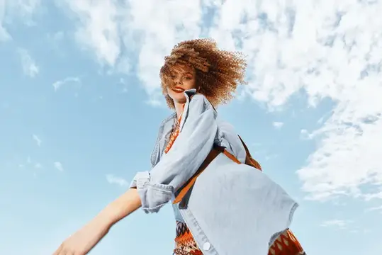 Кудрявая девушка в джинсовой куртке улыбается на фоне неба