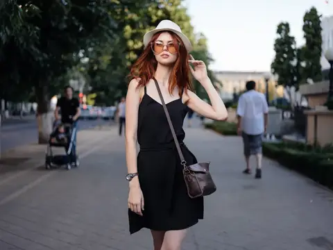 Женщина в черном платье и белой шляпе с сумкой на плече стоит на улице
