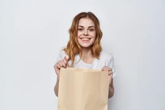 Улыбающаяся рыжеволосая девушка держит крафтовый пакет на белом фоне