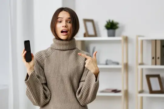 Удивлённая девушка в свитере показывает на телефон