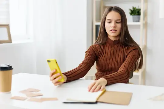 Молодая девушка с жёлтым смартфоном за рабочим столом с блокнотом