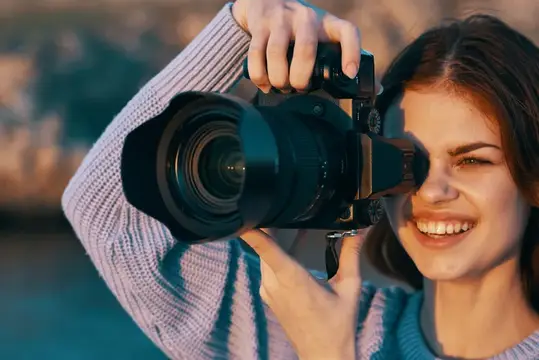 Улыбающаяся девушка смотрит в видоискатель фотокамеры на закате