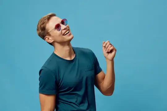 Счастливый молодой человек в повседневной синей футболке и разноцветных солнцезащитных очках, уверенно улыбается и смотрит в сторону, энергичная и беззаботная атмосфера, сплошной ярко-голубой фон…