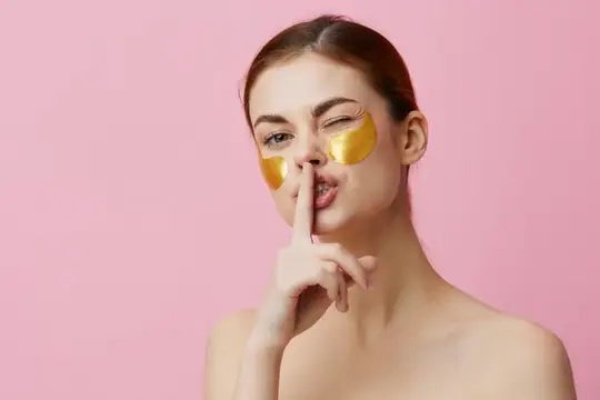 Молодая женщина с золотыми патчами под глазами шутливо подносит палец к губам на розовом фоне