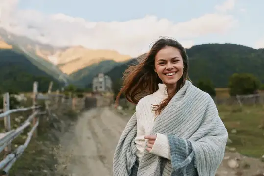 Портрет на открытом воздухе: улыбающаяся женщина на фоне горного пейзажа, в уютном свитере и шали, расслабленный момент жизни среди широкого сельского ландшафта