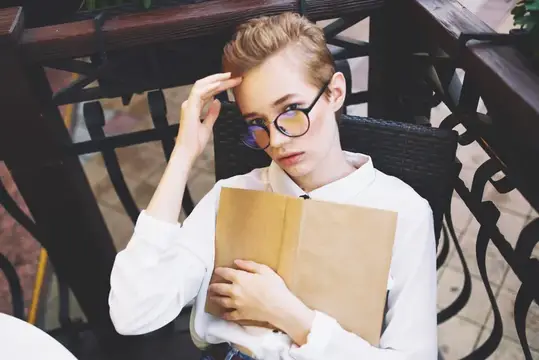 Светловолосая девушка в очках с книгой задумчиво смотрит в сторону на террасе