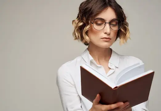 Молодая женщина с волнистыми волосами и в очках читает книгу
