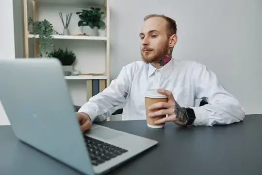 Молодой мужчина с татуировками работает за ноутбуком с кофе
