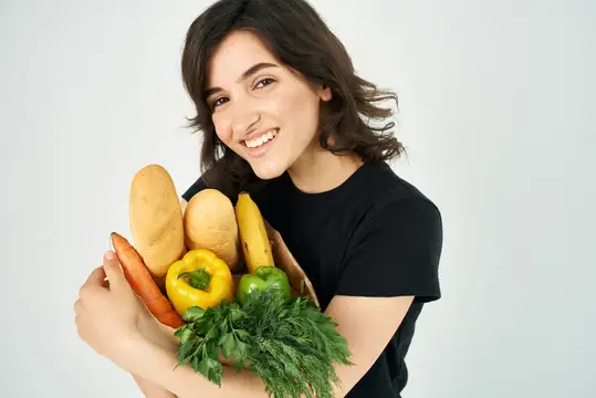 Улыбающаяся темноволосая девушка держит пакет с хлебом и овощами
