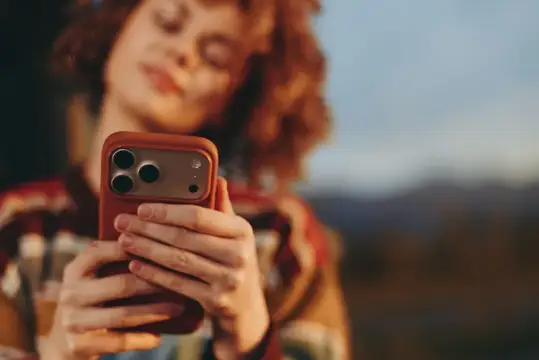 Рыжеволосая девушка держит смартфон в красном чехле на улице